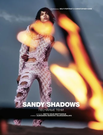 Fashion editorial shot of a model in a lace outfit, posing against a backdrop of a cloudy sky, with a blurred bonfire effect.