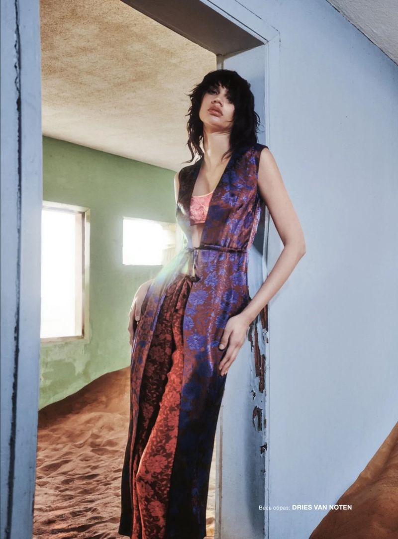 Woman in a patterned sleeveless dress standing in a doorway against an indoor desert scene, illuminated by soft, natural light.
