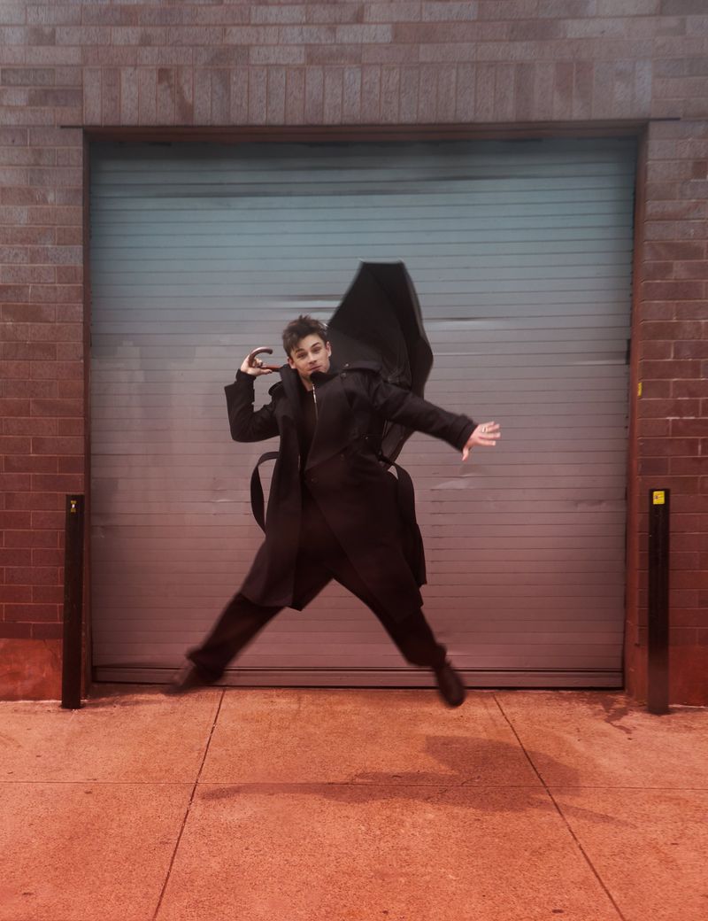 Young adult leaps in front of garage door outdoors. Wearing dark, long-sleeved coat, they hold a closed, black umbrella in hand.