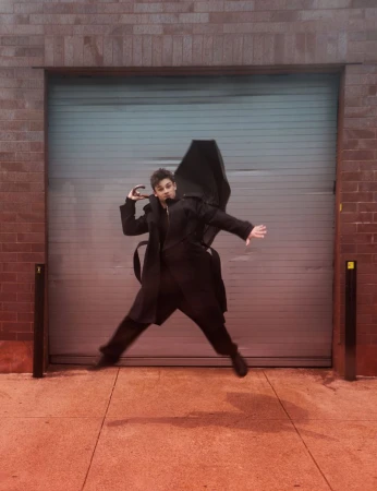 Young adult leaps in front of garage door outdoors. Wearing dark, long-sleeved coat, they hold a closed, black umbrella in hand.