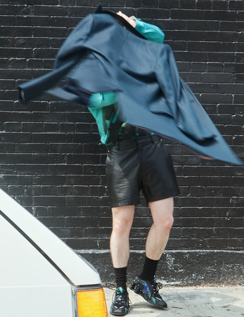Person in leather shorts and iridescent sneakers throwing a blazer in front of a black brick wall with a partially visible white vehicle.