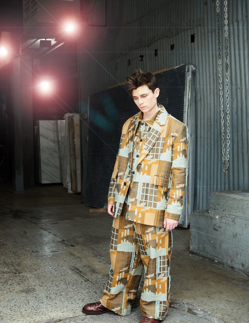 Adult male fashion model wears patterned suit, standing in industrial environment, with moody lighting and urban setting for editorial style shot.
