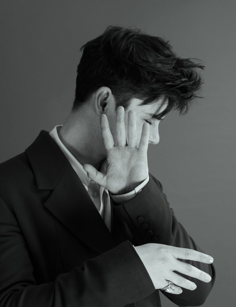 Monochrome portrait of a man in a suit with dark hair covering part of his face with his hand and wearing a ring, against a solid grey backdrop.
