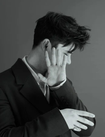 Monochrome portrait of a man in a suit with dark hair covering part of his face with his hand and wearing a ring, against a solid grey backdrop.