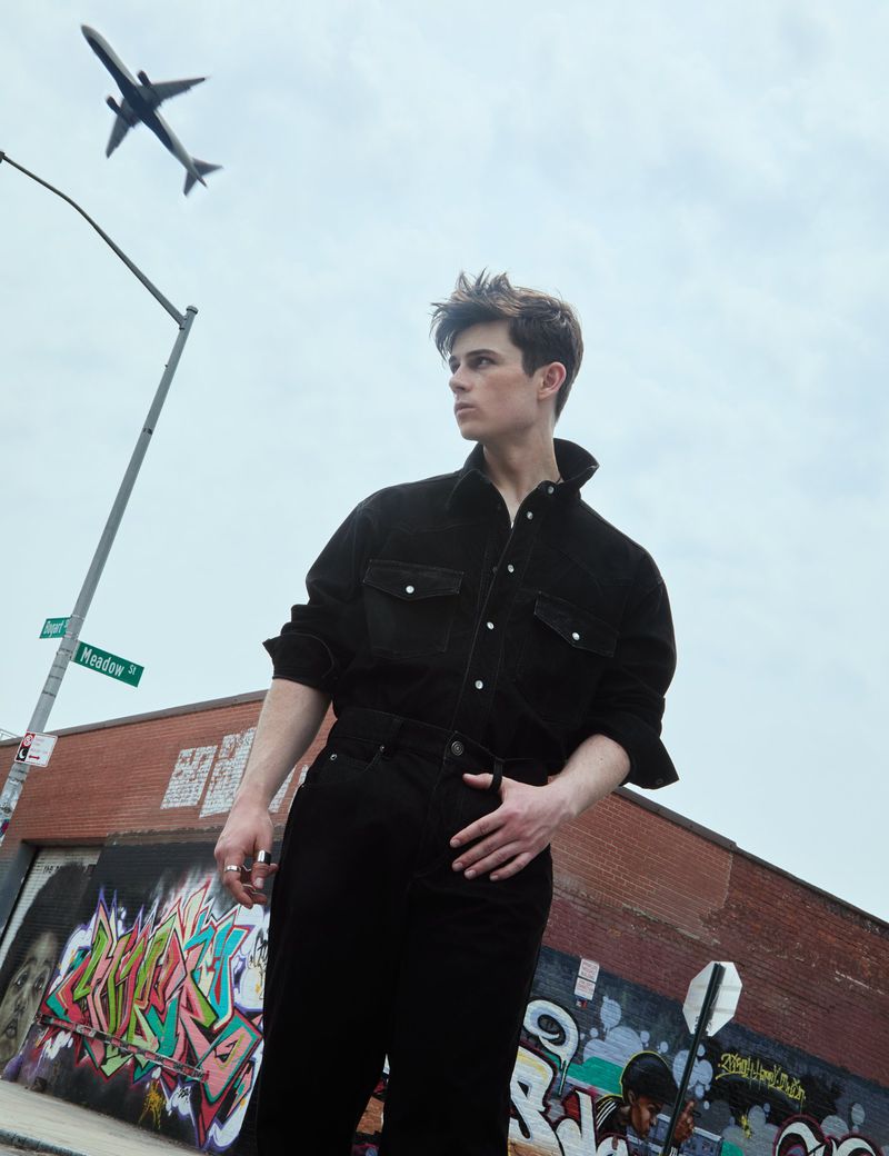 Portrait of a young adult male in dark clothing standing in an urban setting next to graffiti art and a building under an airplane.