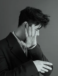 Portrait of Ashby Gentry wearing a dark suit jacket over a white collared shirt, holding up a hand to shield his face.