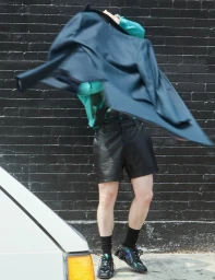 Ashby Gentry flinging a blue jacket over his head wearing a turquoise shirt and black leather shorts against a dark brick wall.