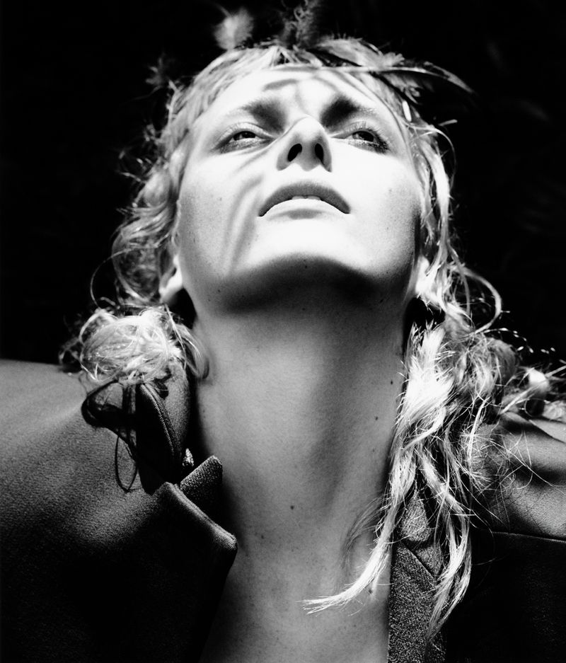 Striking black and white portrait of a fair-skinned adult female looking upwards, adorned with feathers, set against a dark backdrop.