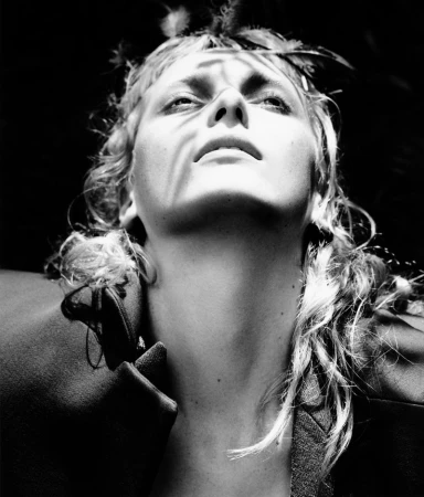Striking black and white portrait of a fair-skinned adult female looking upwards, adorned with feathers, set against a dark backdrop.