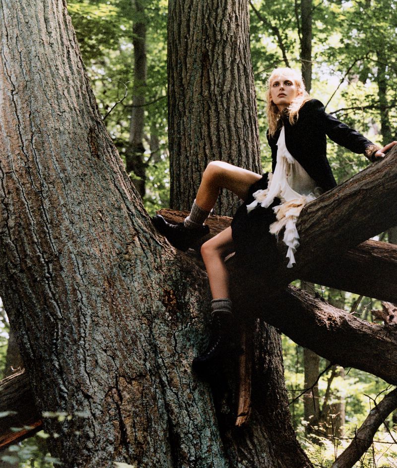 Woman in black jacket and skirt sits on a thick tree branch in a dense forest. Greenery fills the background, creating a natural, woodsy atmosphere.