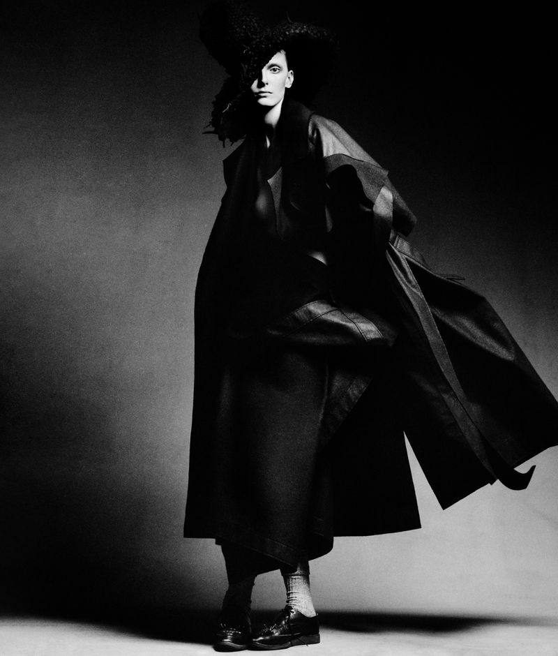 Monochrome fashion shot of a person in a dramatic black hat and flowing coat, posing in a studio setting with dark, textured background.