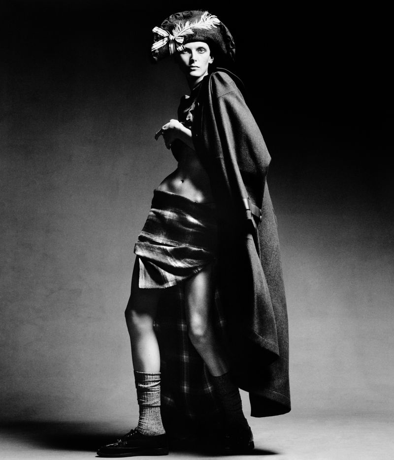 A fashion shot in black and white features a slender adult female model wearing a cap, long cape, and draped skirt over a gray backdrop.