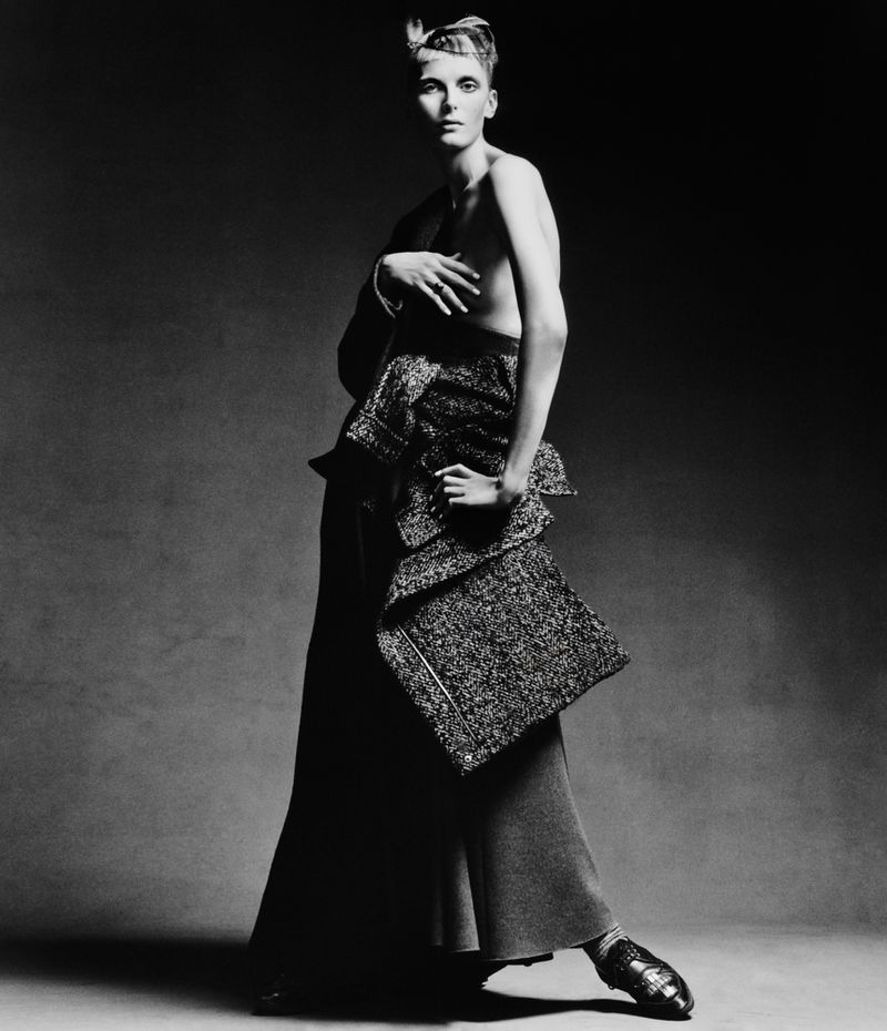 A fashion portrait in black and white featuring a female model with short hair posing in an elaborate top and long skirt in a studio setting.