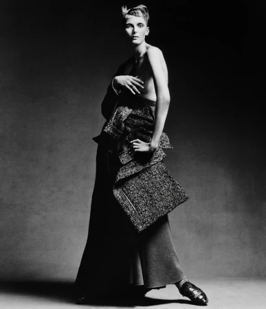 A fashion portrait in black and white featuring a female model with short hair posing in an elaborate top and long skirt in a studio setting.