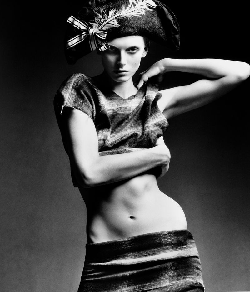 Monochrome studio shot of a slim adult female wearing a striped top, skirt, and feathered hat, posing with hand raised to her shoulder.