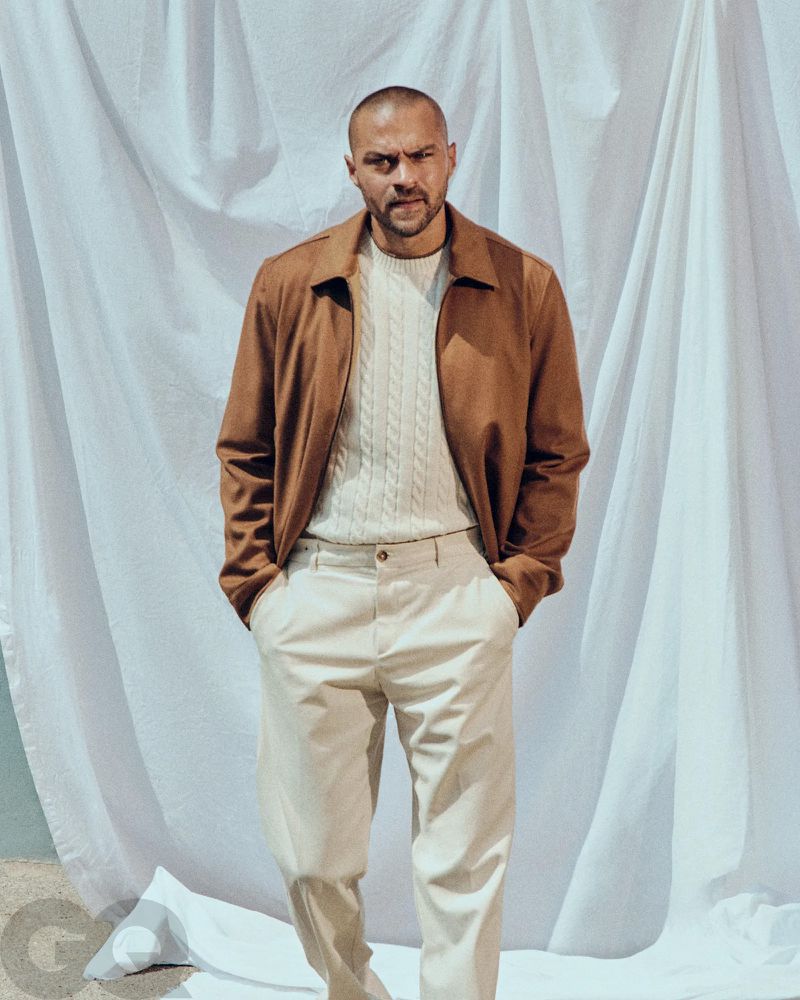 Adult male in a medium shot wearing a brown jacket and white pants stands against a white backdrop. GQ logo visible in lower left corner.