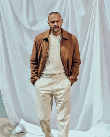 Adult male in a medium shot wearing a brown jacket and white pants stands against a white backdrop. GQ logo visible in lower left corner.