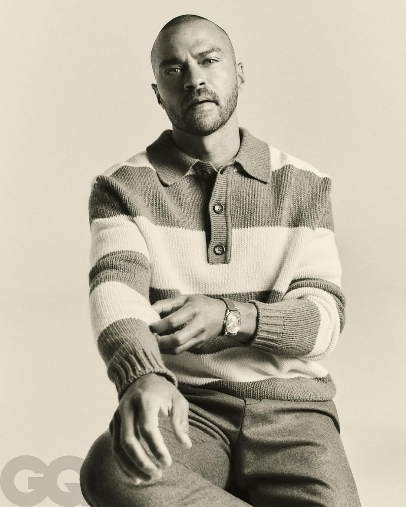 Jesse Williams poses in a black and white medium studio shot, wearing a striped sweater, dress pants, and a wristwatch.