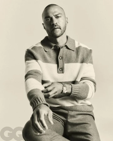 Jesse Williams poses in a black and white medium studio shot, wearing a striped sweater, dress pants, and a wristwatch.