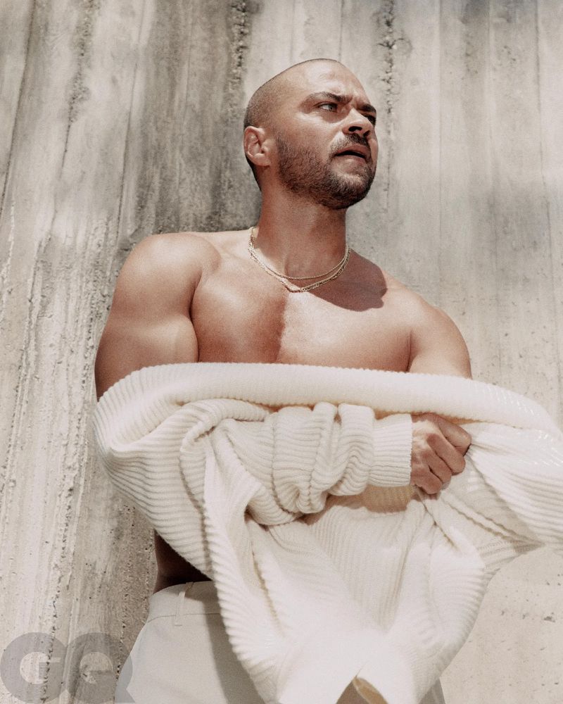 Jesse Williams poses shirtless in off-white pants, holding a textured jacket against a textured wall for a fashion editorial shoot.
