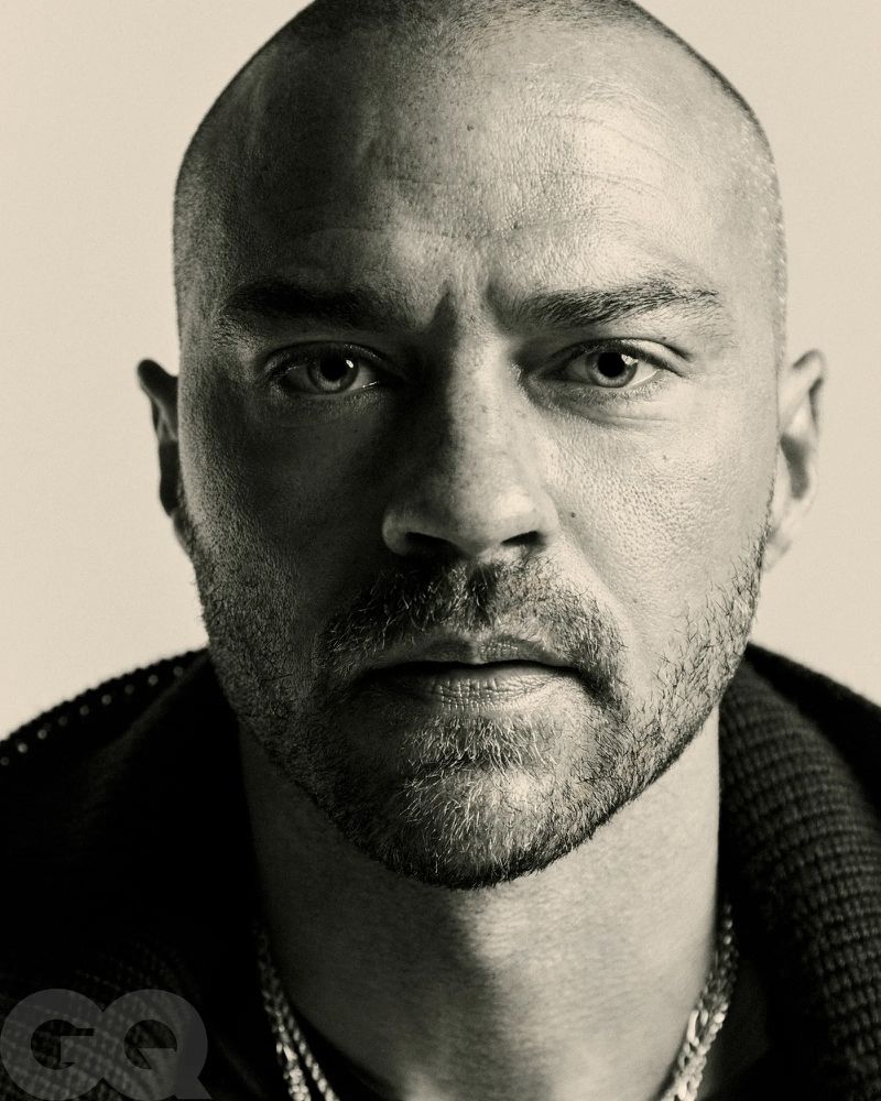Close-up studio portrait of a bald adult man with a beard. He is wearing a necklace and dark jacket, looking intently at the camera.