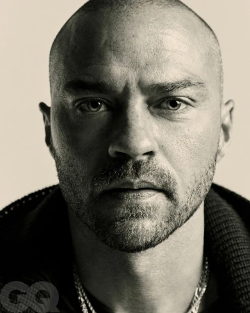 Close-up studio portrait of a bald adult man with a beard. He is wearing a necklace and dark jacket, looking intently at the camera.