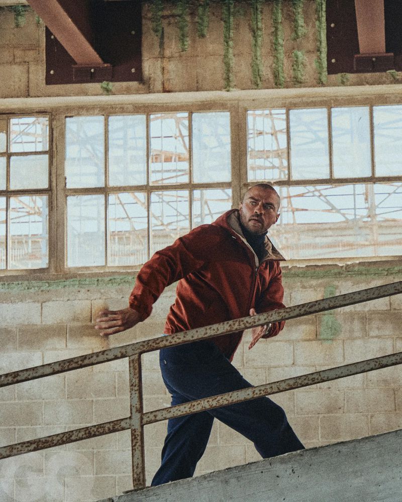 Adult man in an orange jacket climbs stairs in an abandoned building with large windows and concrete walls with some vegetation.