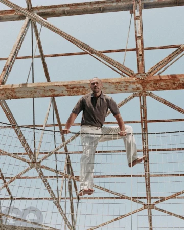 Adult male, barefoot, poses on a rusty metal structure against a pale blue sky in an outdoor setting. He is wearing a short-sleeved shirt and pants.