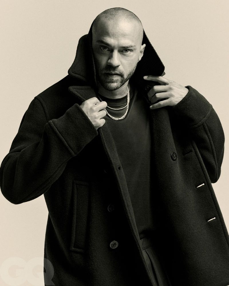 Jesse Williams poses in a studio portrait, wearing a black peacoat and silver chain against a neutral backdrop, exuding a cool, confident vibe.