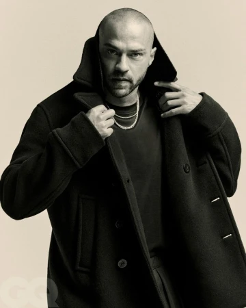 Jesse Williams poses in a studio portrait, wearing a black peacoat and silver chain against a neutral backdrop, exuding a cool, confident vibe.