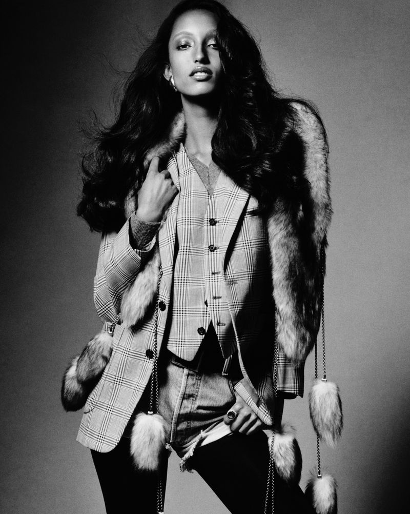 A fashionable adult female model in a plaid blazer and fur stole, photographed in a studio with a gray background.