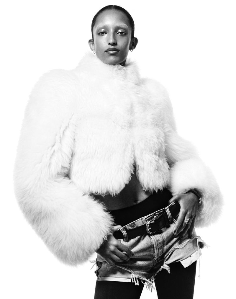 Black and white studio shot of a slender adult woman wearing a white faux fur jacket, denim shorts, and tights against a white backdrop.