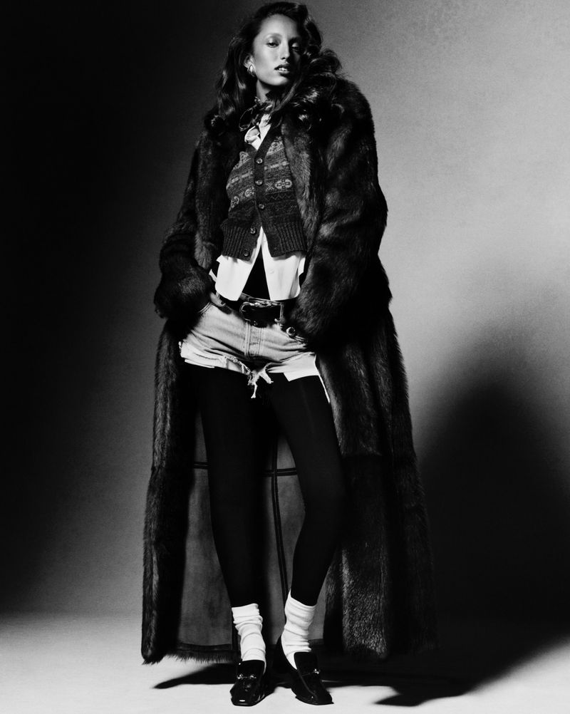 Fashion model in faux fur coat, cardigan, and denim shorts, posing in a photo studio with black and white grading and medium shot.