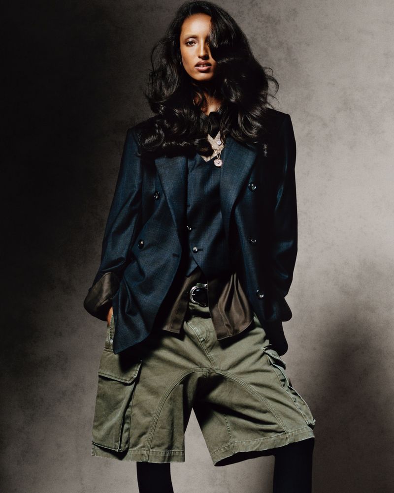 A woman poses in an unbuttoned blazer, green cargo shorts, and leggings against a mottled grey backdrop in a studio setting.