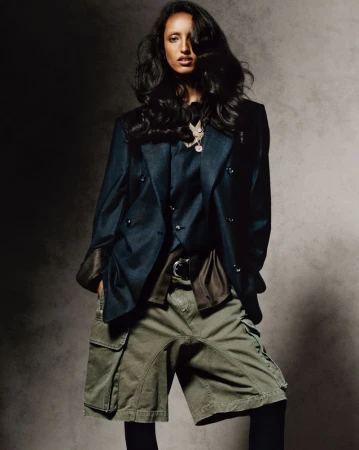 A woman poses in an unbuttoned blazer, green cargo shorts, and leggings against a mottled grey backdrop in a studio setting.