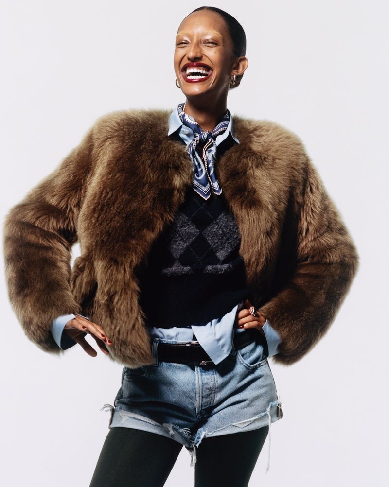 Smiling woman wearing fur coat, denim shorts, black tights, and boots against a white backdrop. Fashion and portrait photography.