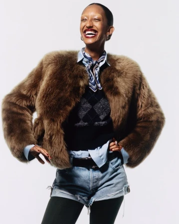 Smiling woman wearing fur coat, denim shorts, black tights, and boots against a white backdrop. Fashion and portrait photography.