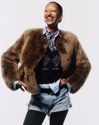 Model Mona Tougaard laughing in a brown fur jacket, argyle sweater vest, denim cutoffs, and black tights against a white background.