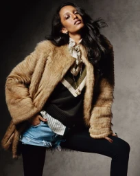 Model Mona Tougaard poses in a camel fur coat, layered knitwear, and denim shorts against a textured grey studio backdrop.