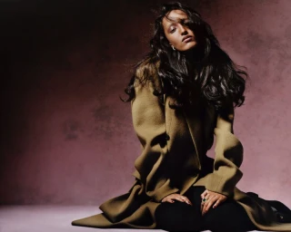 Model Mona Tougaard seated in an oversized brown wool coat against a textured mauve studio backdrop.