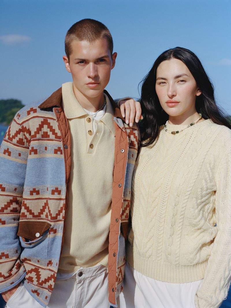 Two young adults, a man and woman, model knitwear against a background of blue sky and a distant wooded shoreline in natural light.
