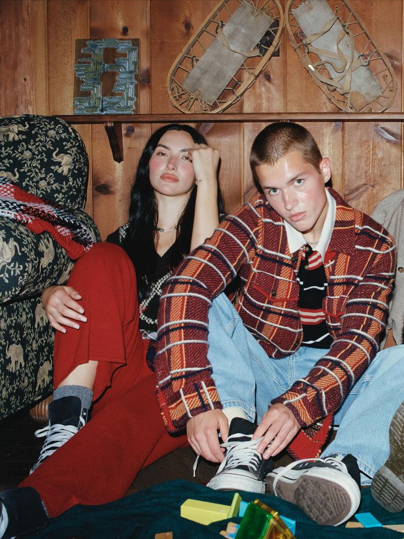 Two young adults sit on a floor next to a wall with decor, posing in a medium shot. They wear street-style casual clothing with bold plaid patterns.