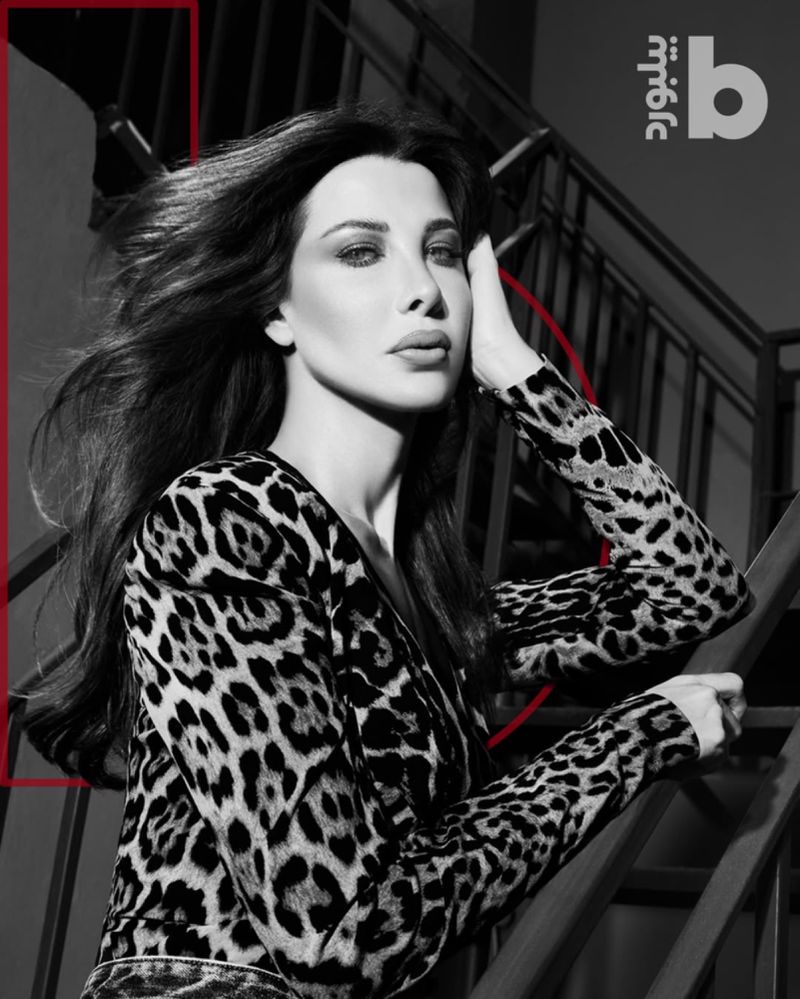 Portrait of an adult female with long wavy dark hair, wearing an animal print top, posing indoors on a staircase with grey tones.
