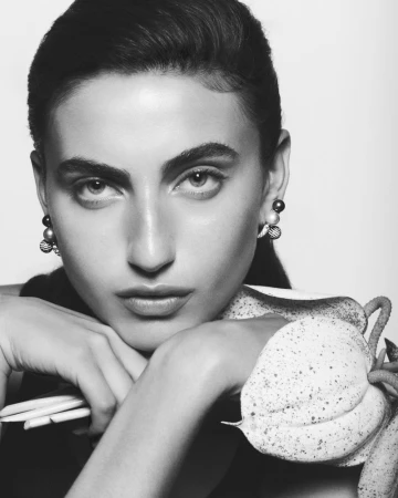 Striking monochrome portrait of a woman with sleek dark hair, complemented by pearl earrings, gently resting her chin on her hands.