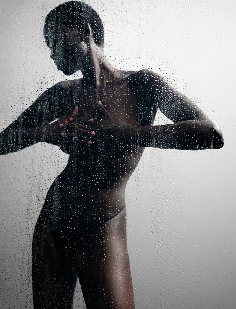 Studio portrait of a slender African American woman posing behind a textured glass surface, with water droplets, against a grey background.