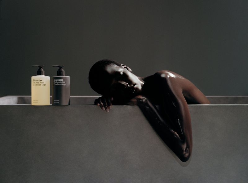 Person in bathtub with two bottles of body lotion. Dark grey studio shot with contrasting tones and textures, highlighting skincare products.