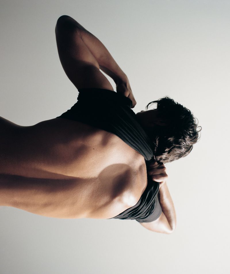 Close-up studio shot of an adult male, seen from the back, removing his black t-shirt in a brightly lit studio environment.