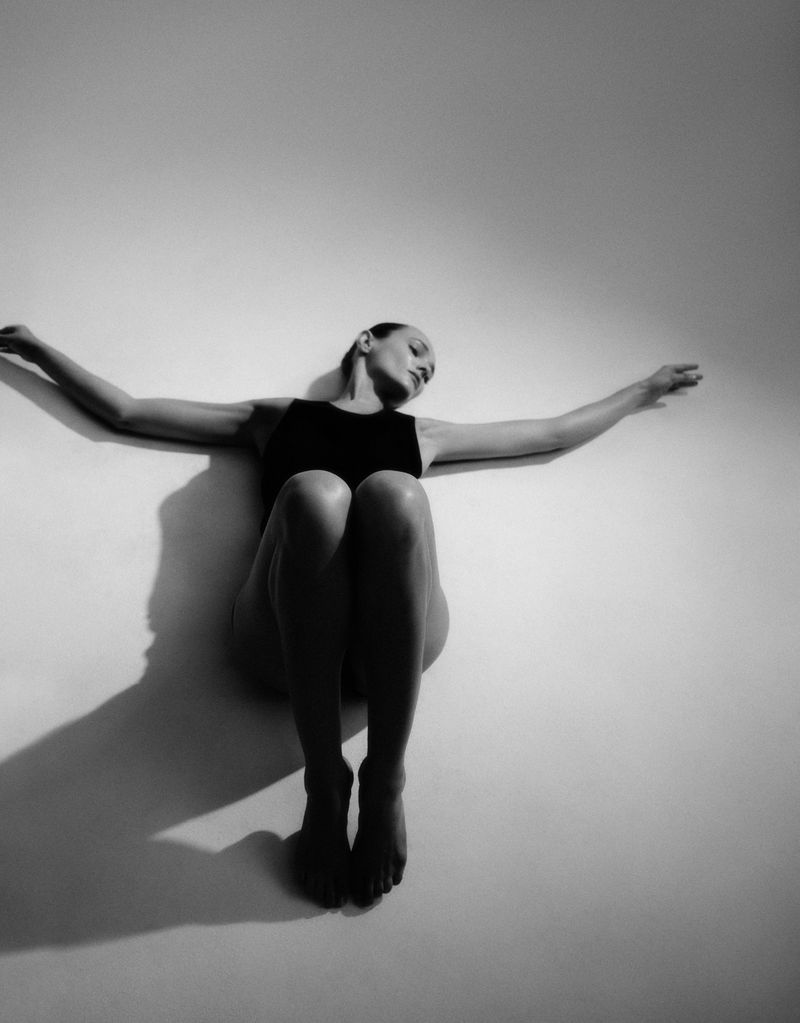 Artistic black and white studio portrait of an adult woman in a dark swimsuit, stretching her arms to the sides on a white wall.
