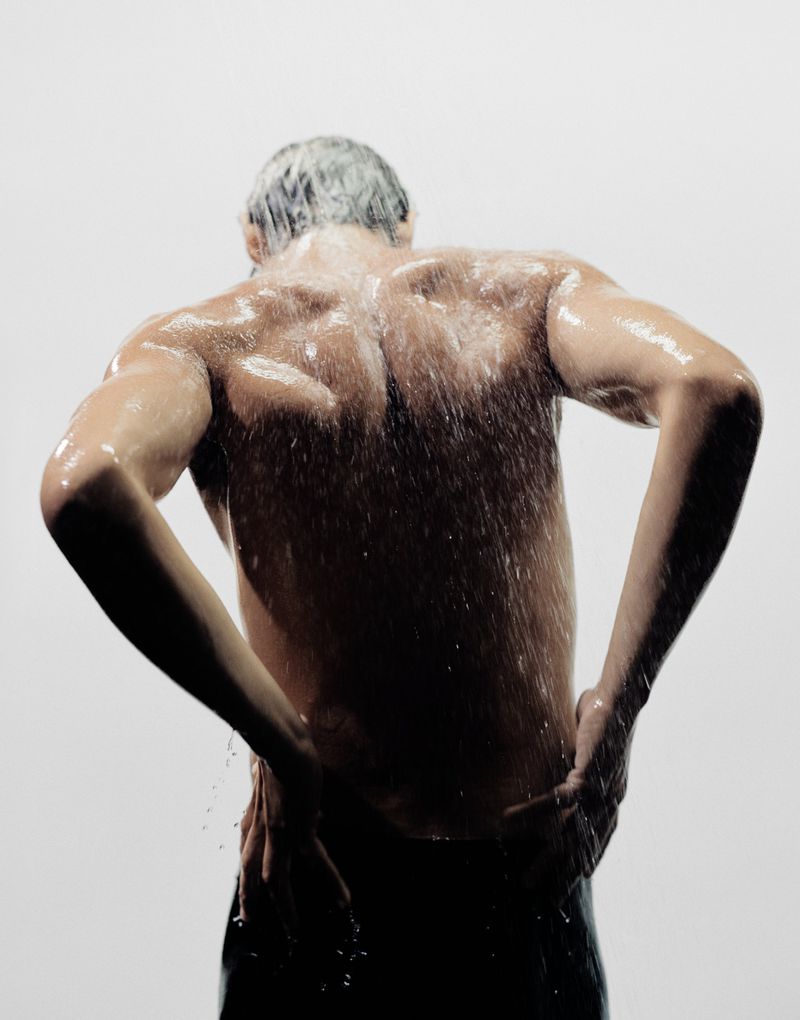 Rear view of a muscular man with water cascading down his back. The man is holding his hips with his hands in a bright studio.