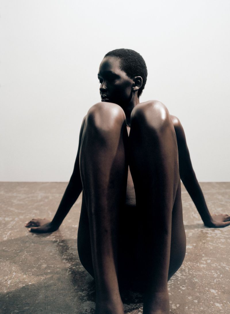 Nude African woman sitting on the floor with her knees up, looking off to the side in a minimalist photo studio setting with warm light.
